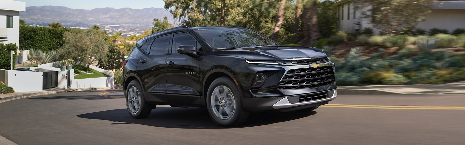 Black 2025 Chevrolet Blazer SUV driving on a road surrounded by modern homes and greenery, showcasing its sleek exterior design and stylish front grille.
