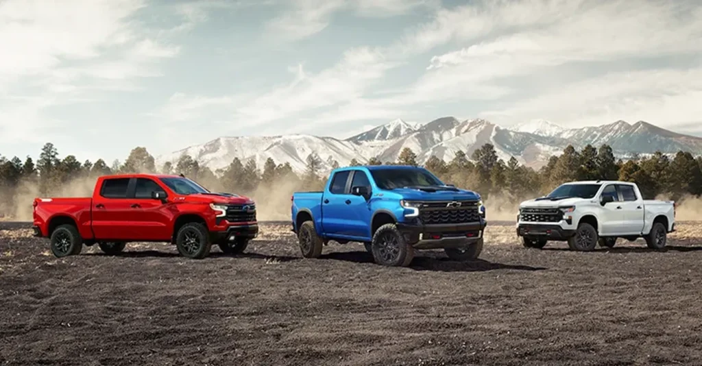 The image displays three 2026 Chevrolet Silverado trucks in vibrant colors: red, blue, and white, set against a rugged, mountainous backdrop. These trucks, with their off-road tires, are positioned on dirt terrain, emphasizing their powerful and adventurous design.