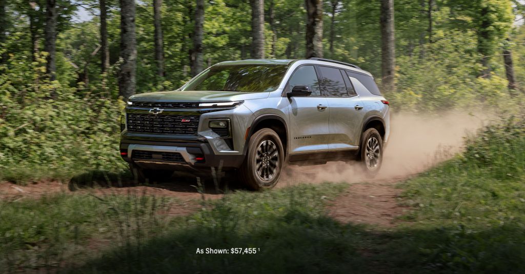 A silver 2026 Chevy Traverse driving off-road on a wooded dirt trail.