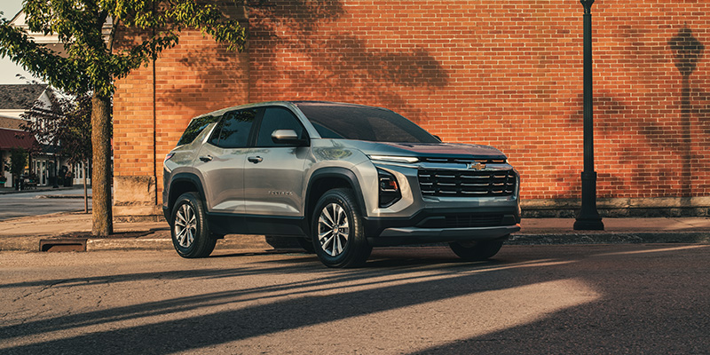 Equinox parked by a brick wall on a sunny street, under a tree with dappled shadows. The scene feels calm and suburban.